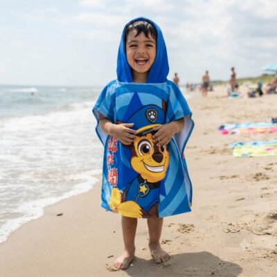 Toddler on the beach wearing a blue Paw Patrol towel and hood, smiling at the camera.
