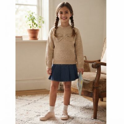 Young girl standing indoors in a beige sweater with a bow, a denim pleated skirt, white socks and pink sneakers, holding a book.