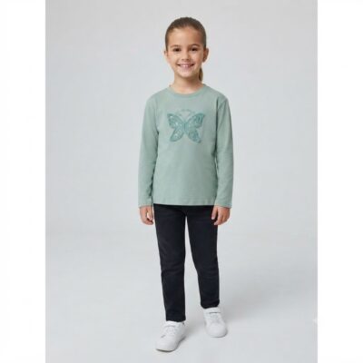 Smiling girl in a mint-green long-sleeve shirt with a glitter butterfly graphic, dark jeans, and white sneakers, standing in a neutral studio.
