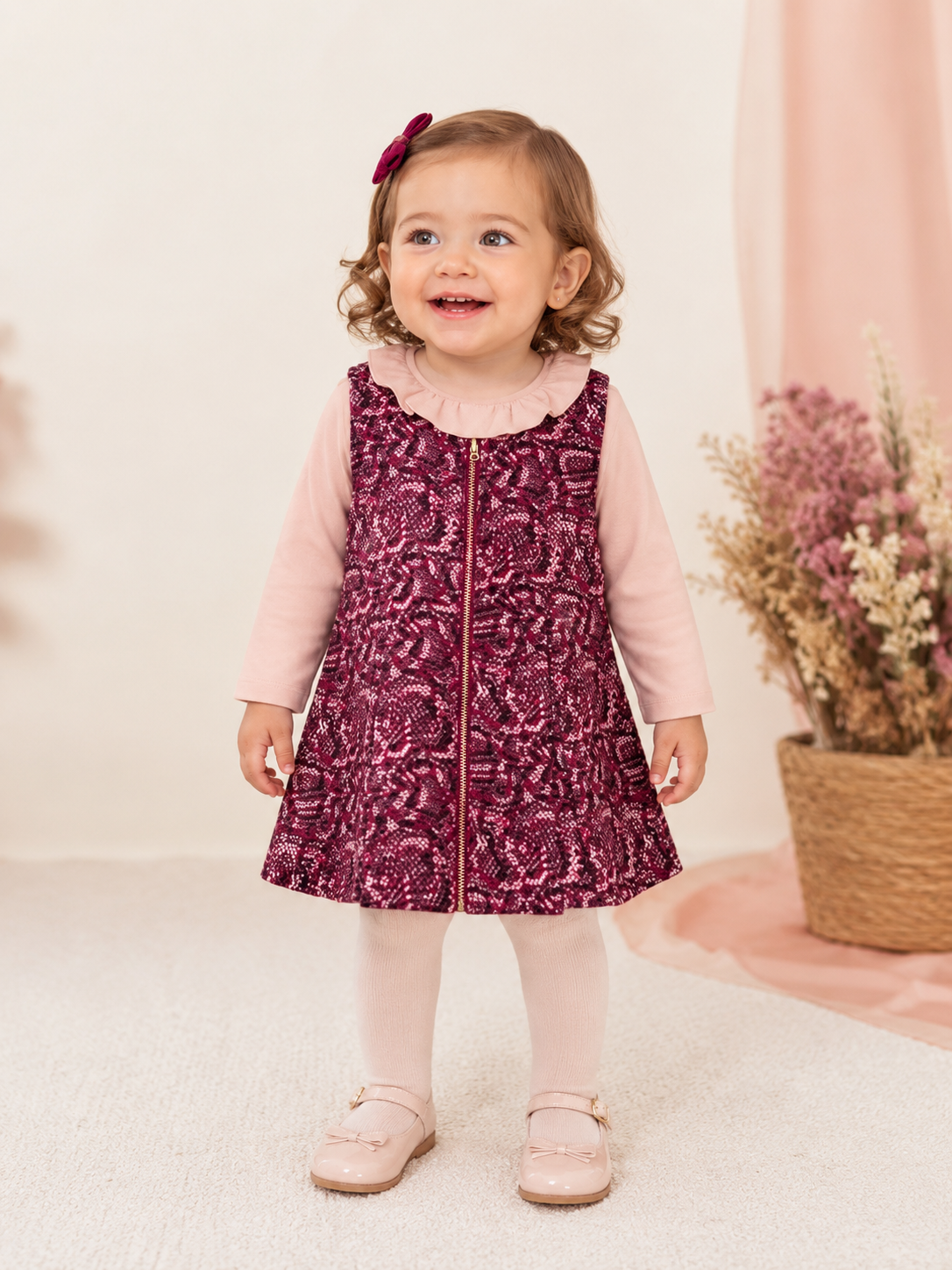 e0cb1d41-ec2b-417c-b279-660179426bd5 - Da-Da Kids Smiling toddler girl stands indoors in a pink top and burgundy patterned dress with a gold zipper, wearing a pink bow in her hair and pink shoes with tights. Background shows dried flowers in a basket. - Alternative 2: Little girl posing in a pink long-sleeve shirt under a burgundy patterned dress, with a bow in her hair and pale-pink shoes. - Alternative 3: Adorable toddler wearing pastel pink tights and patent shoes, standing on a light carpet in a soft-toned room with a floral backdrop.