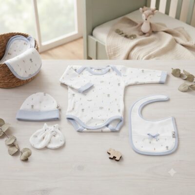 Baby clothing set laid out on a light wooden table: white and blue patterned onesie, hat, mittens, bib, and blanket in a nursery.