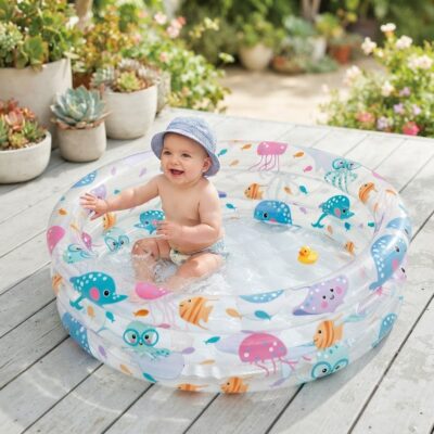 e0545051-f69b-4474-8508-9128aa4065b7 - Da-Da Kids Baby sitting in a colorful inflatable kiddie pool on a wooden deck, wearing a striped hat and smiling, with potted succulents in the background.