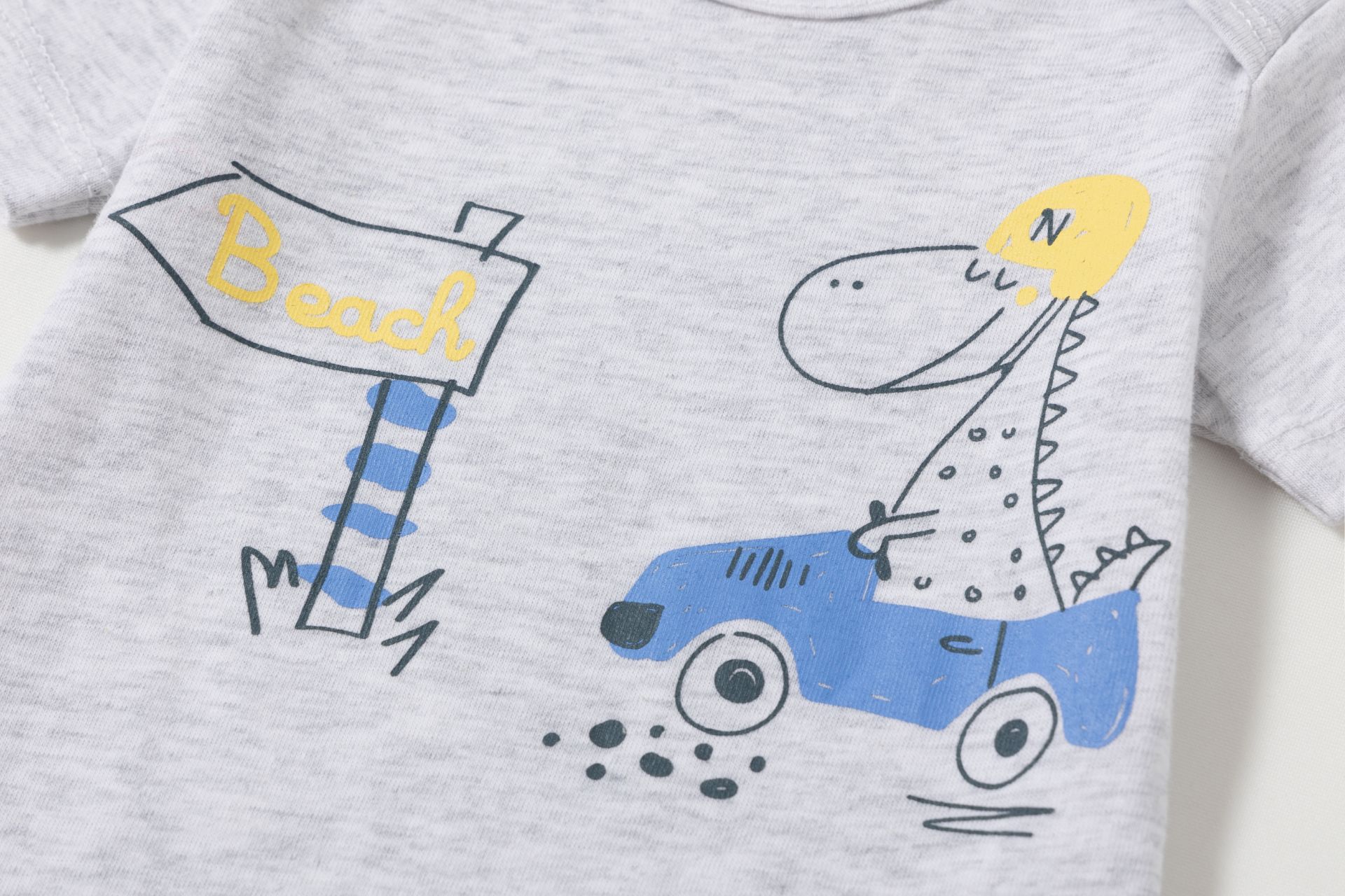 0021072_bodies-3-a-24-meses - Da-Da Kids Gray T‑shirt featuring a cartoon dinosaur driving a blue car with a yellow helmet and a sign that reads 'Beach' in yellow letters.