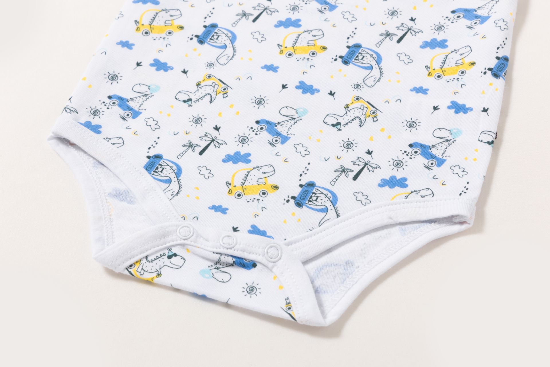 0021075_bodies-3-a-24-meses - Da-Da Kids Close-up of a white baby onesie fabric with a playful print of blue and yellow dinosaurs, cars, palm trees, and clouds.