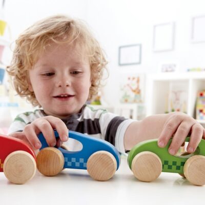 0fcb4434-08ef-439b-980c-23ab140ea8f8 - Da-Da Kids Young child plays with three wooden toy cars (red, blue, green) on a white table in a bright room.