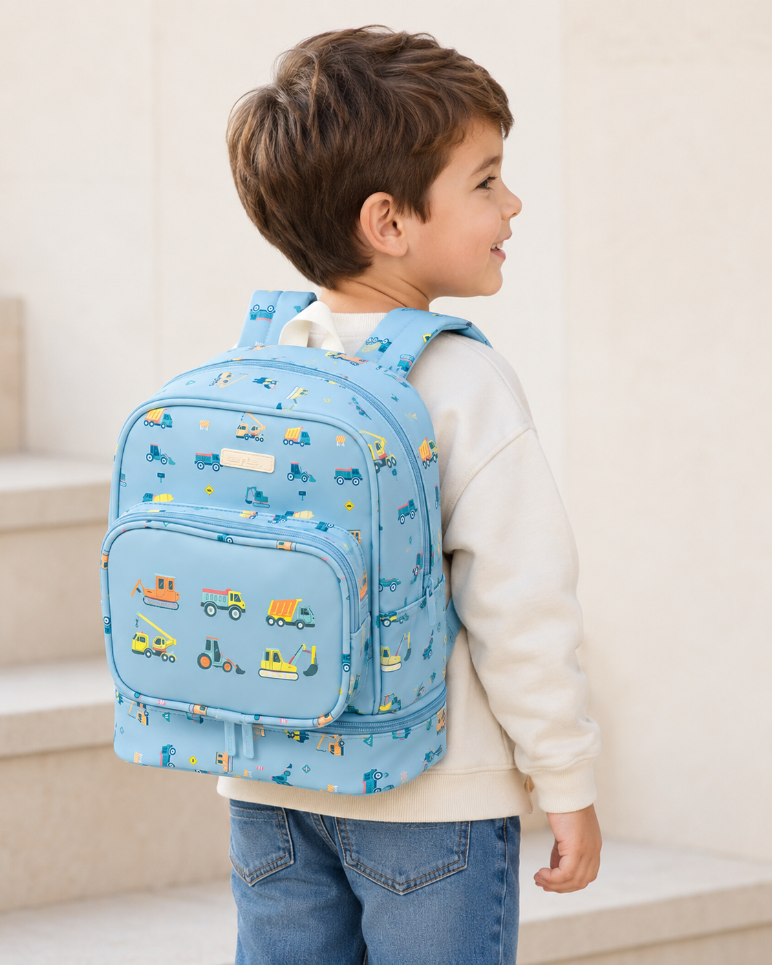 3a91dd8b-a12e-463f-b130-e5b135c2a269 - Da-Da Kids Young boy in a beige sweater wearing a blue backpack with construction vehicle pattern, standing on steps.