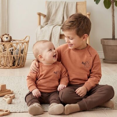 Two brothers wearing matching orange outfits sit on a rug, smiling at each other in a cozy playroom.