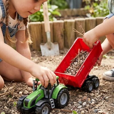Two children play in a garden with a green toy tractor pulling a red wagon filled with rocks and twigs.