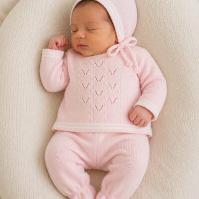 Sleeping newborn in a soft pink knit outfit on a cream blanket, wearing a matching cap with a bow.