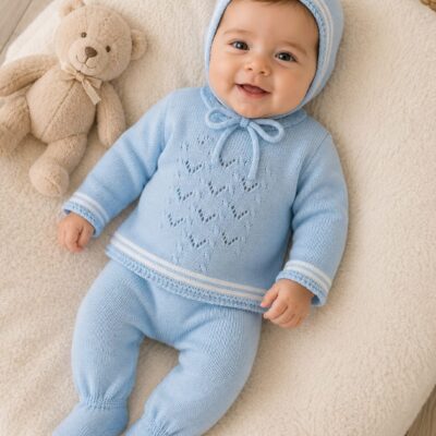 Smiling baby in a light blue knit outfit and bonnet lying on a cream blanket beside a plush teddy bear.