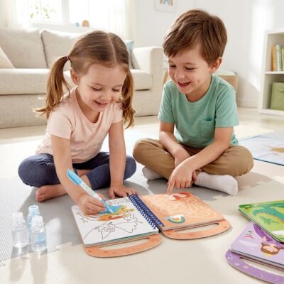 beebb19a-4667-4764-8b25-4f9784bb473d - Da-Da Kids Two kids sit on a soft play mat in a bright living room, coloring in spiral-bound activity books with markers.