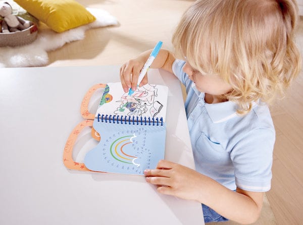 hape-toys-water-doodle-pad-1227737219 - Da-Da Kids Blonde toddler sitting at a white table, coloring a small rainbow-themed activity book with a blue marker.