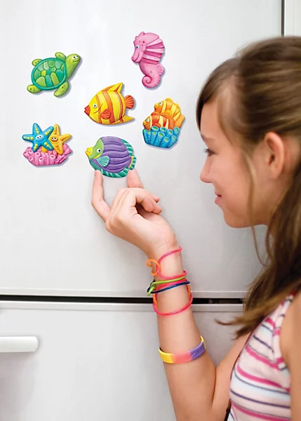 mould-paint-craft-kit-sealife~4893156035110_04c_MP - Da-Da Kids Girl wearing colorful bracelets places magnetic sea-creature magnets on a white refrigerator door, including a turtle, fish, seahorse, starfish, and shells.
