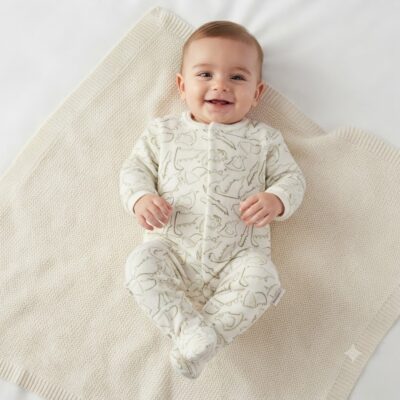 Smiling baby lying on a cream knit blanket in a white patterned onesie.