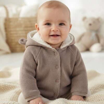 fc17eba2-0c49-4f4d-8bfa-03caaf2cc055 - Da-Da Kids Smiling baby sitting on a cream knitted blanket, wearing a brown hooded cardigan in a bright, cozy room with a teddy bear in the background.